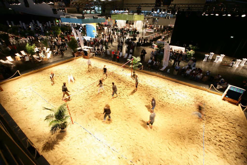 Beachvolleyballfeld in einer Messehalle mit spielenden Personen, leicht verschwommen, Zuschauer stehen drum herum
