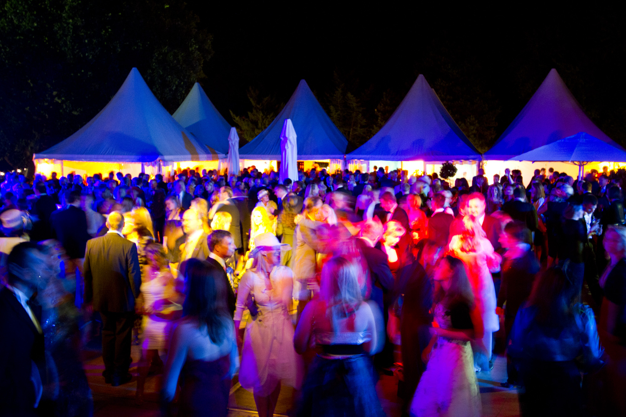 festlich gekleidete Partygäste, tanzen im bunten Scheinwerferlicht vor fünf weißen, kleinen Festzelten