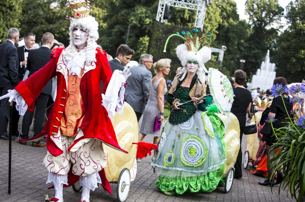 Künstler in barocken Kostümen auf Schnecken-Fahrzeugen beim Kulturprogramm im Stadtpark Hannover. Im Hintergrund mischen sich festlich gekleidete Eventgäste.