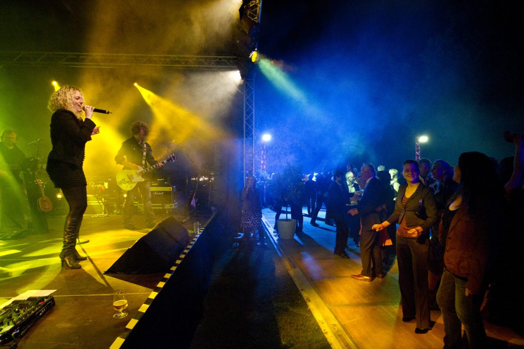 festlich gekleidete Partygäste, tanzen vor einer bunt, erleuchteten Bühne, auf der eine Band mit Sängerin Musik macht.