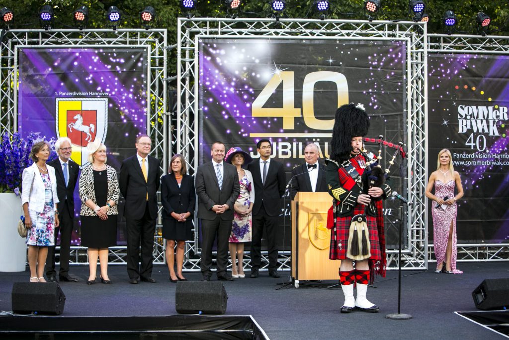 Ein Dudelsackpfeifer in traditionellem Kilt spielt auf einer Bühne bei der Jubiläumsfeier zum 40. Sommerbiwak der 1. Panzerdivision Hannover. Im Hintergrund stehen Ehrengäste, darunter Militär- und Zivilpersonen in formeller Kleidung. Ein Banner mit der Aufschrift '40. JUBILÄUM' und 'SOMMERBIWAK 40' ist zu sehen.